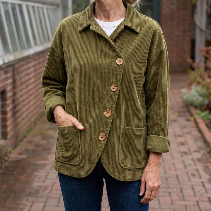Cervinia Collared Jacket — Olive, worn by a woman with silver-blonde hair over a white tee and dark jeans, one hand in the pocket, cropped at the neck, outside a glasshouse on a brick path