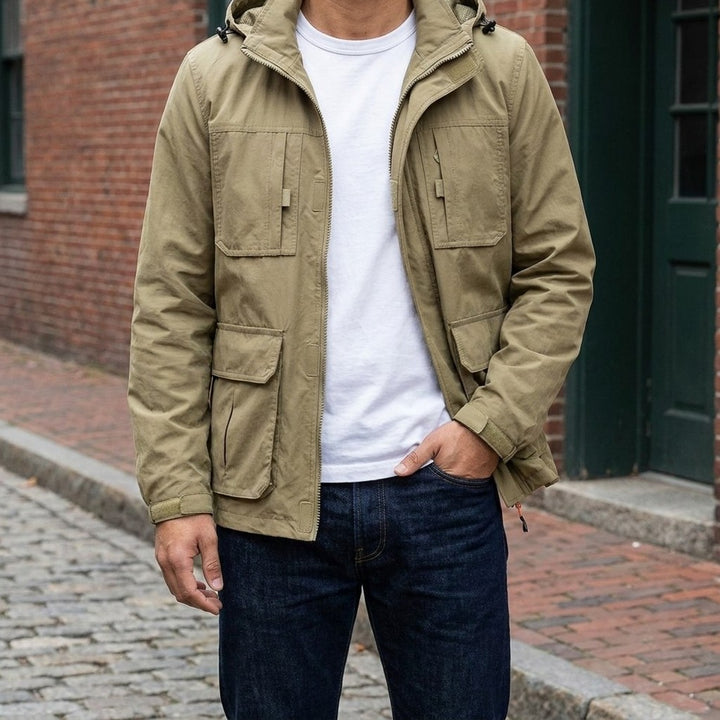 Man wearing the Faenza Hooded Cargo Jacket in Khaki open over a white tee on a brick-lined street