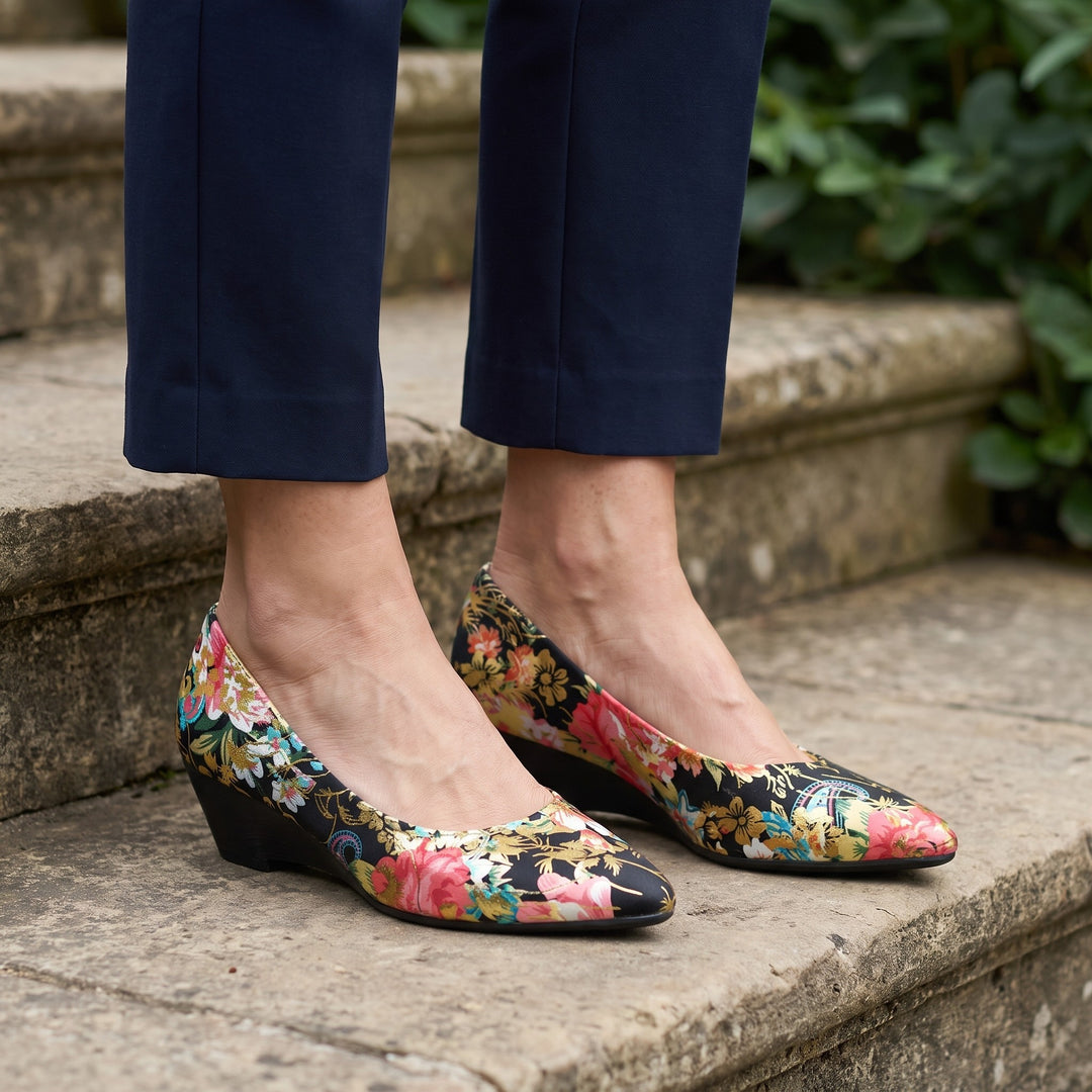 Maiori Pointed-Toe Pumps — Black Floral, worn by a woman in navy cropped trousers, knee down, on stone garden steps