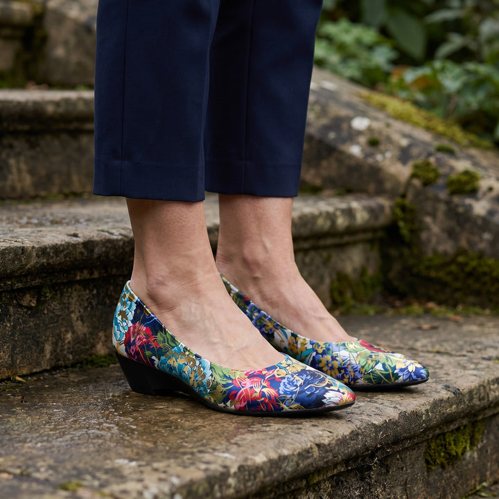 Maiori Pointed-Toe Pumps — Teal Floral, worn by a woman in navy cropped trousers, knee down, on stone garden steps