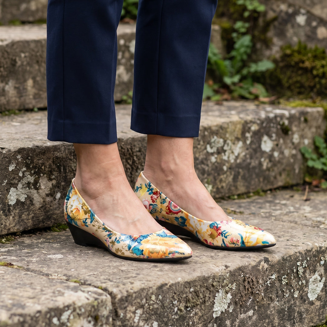 Maiori Pointed-Toe Pumps — Cream Floral, worn by a woman in navy cropped trousers, knee down, on stone garden steps