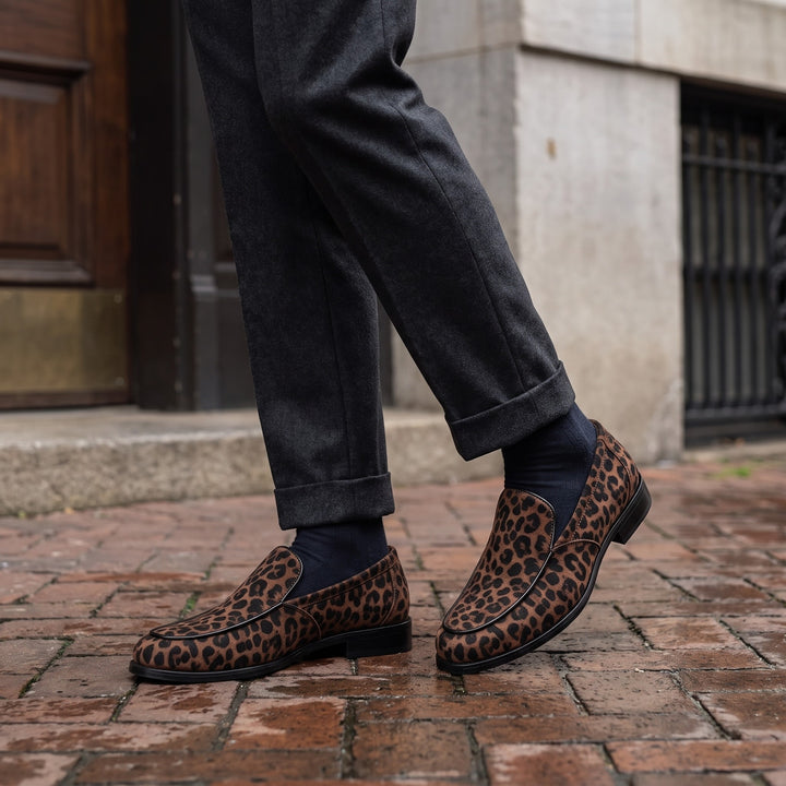 Mantova Suede Loafers — Brown Leopard, worn by a man in charcoal trousers, knee down, on a brick sidewalk outside a historic hotel entrance