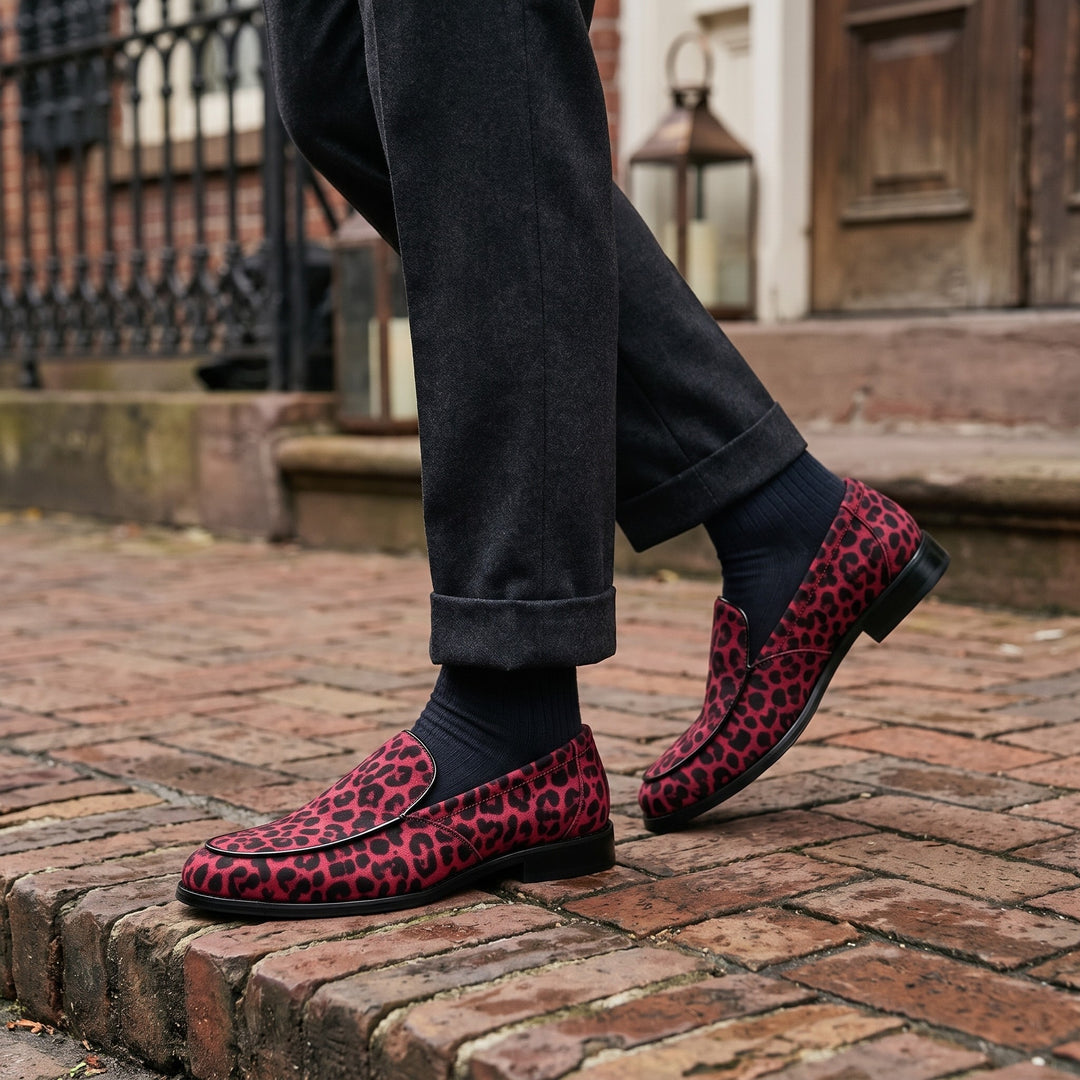 Mantova Suede Loafers — Red Leopard, worn by a man in charcoal trousers, knee down, on a brick sidewalk outside a historic hotel entrance