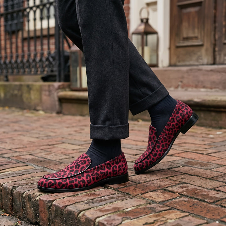 Mantova Suede Loafers — Red Leopard, worn by a man in charcoal trousers, knee down, on a brick sidewalk outside a historic hotel entrance